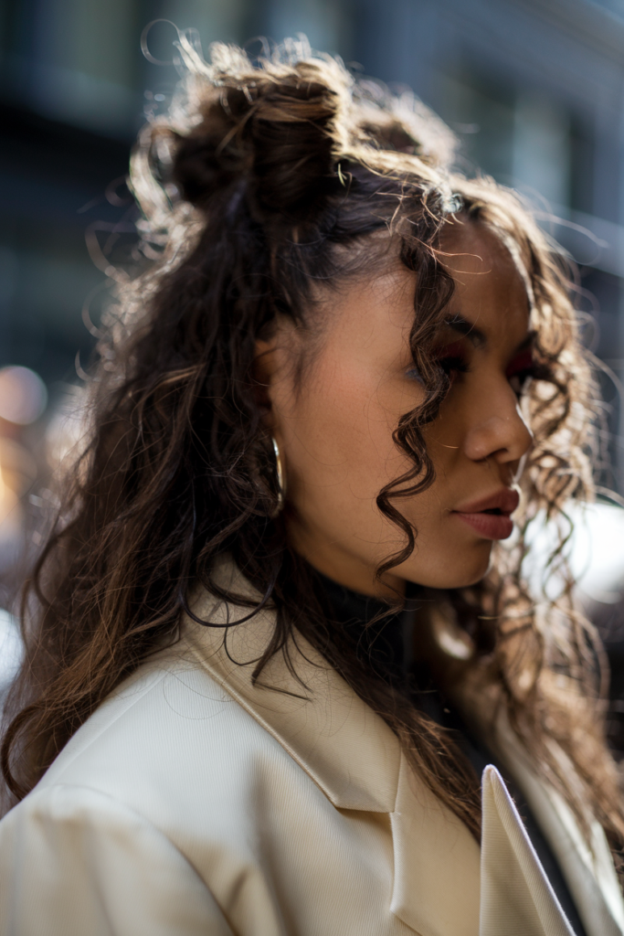 A bright street style photo of a woman with curly space buns wearing an oversized cream blazer. The photo is taken in Soho with natural morning light. The woman has rich melanated skin. The shot is taken with an 85mm lens and has a fashion week aesthetic.